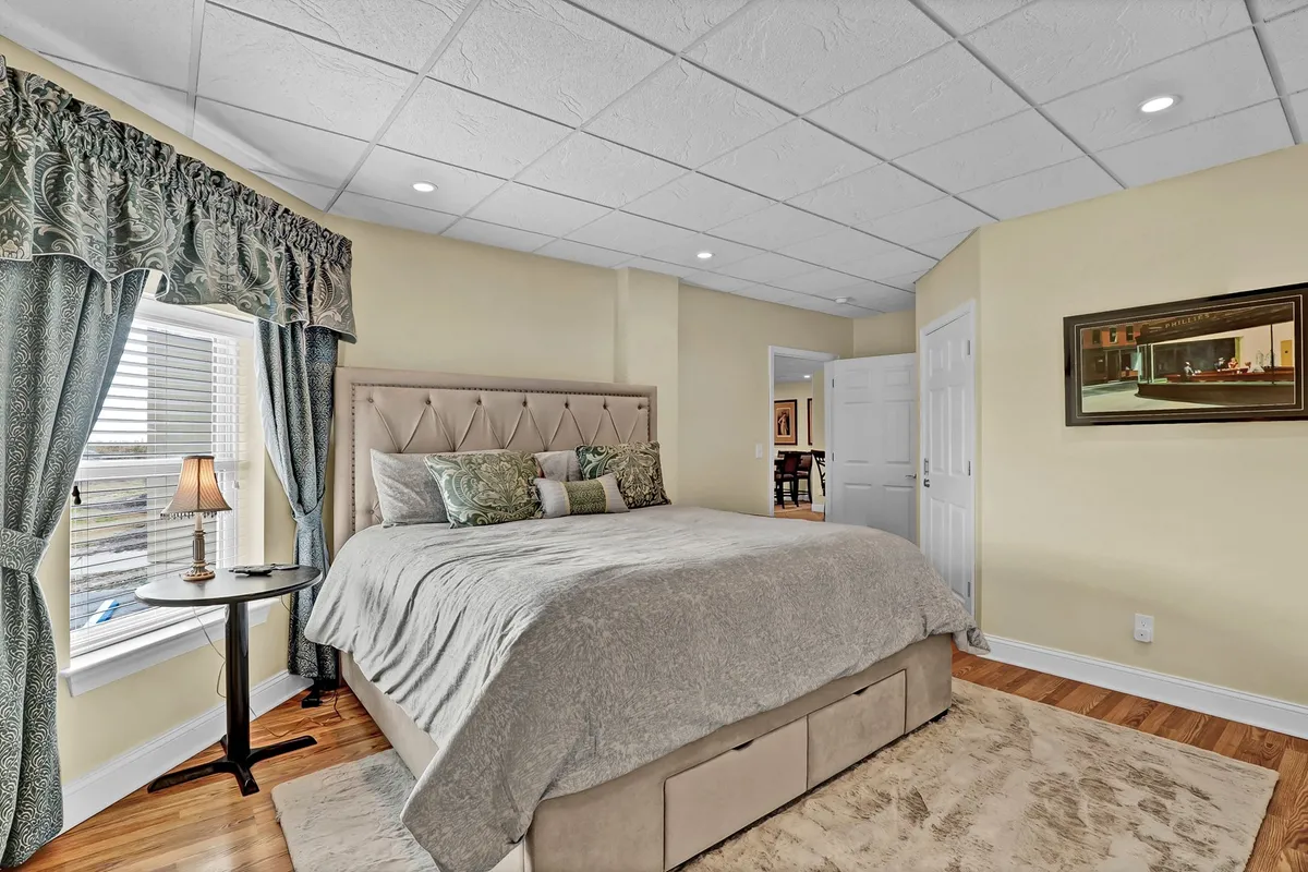 A beautifully styled bedroom featuring a plush bed with inviting linens, flowing curtains softly filtering the natural light, a nightstand with a lamp, and calming wall art. The atmosphere is tranquil and designed for restful relaxation.