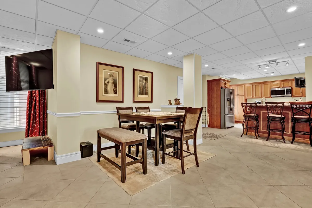 The image features a dining table near a bar area, with framed artwork and a TV in the background. The layout encourages both dining and entertainment in a welcoming environment.