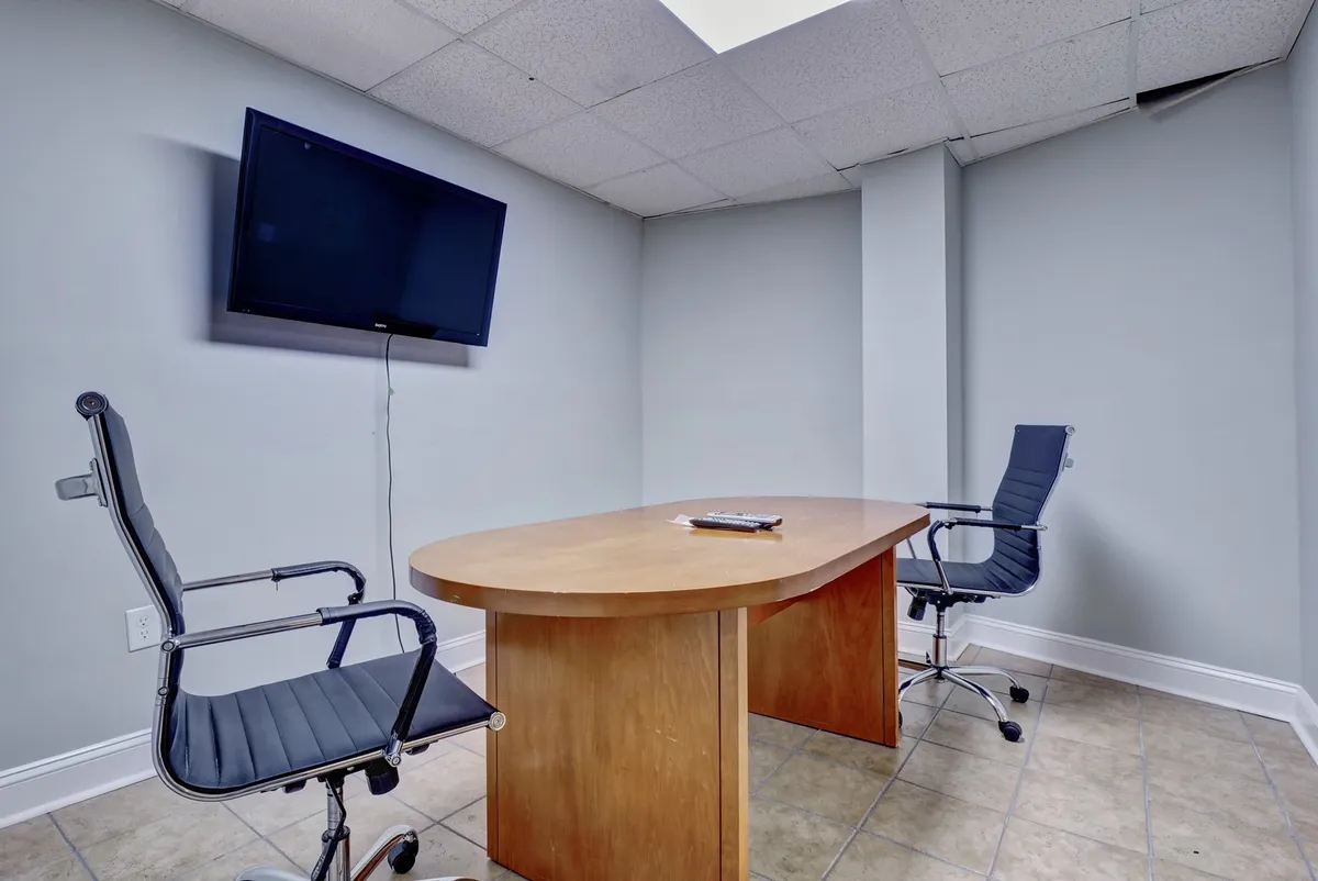 A modern conference room featuring a wooden table, two ergonomic office chairs, and a wall-mounted television, creating a dedicated workspace for meetings, video calls, or focused work sessions.