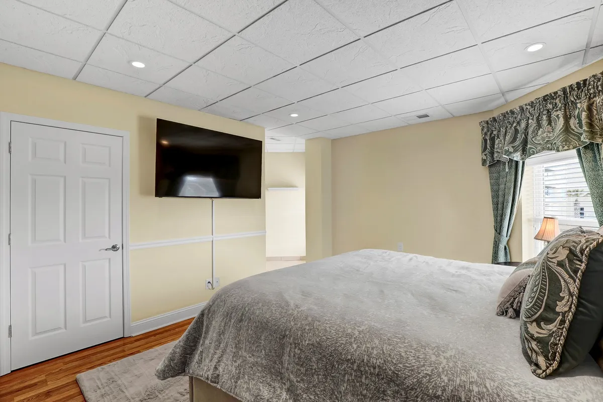 A cozy bedroom with a comfortable bed adorned with soft bedding, elegant window treatments allowing gentle light, and a wall-mounted television for late-night unwinding. The ambiance is inviting and perfect for relaxation.