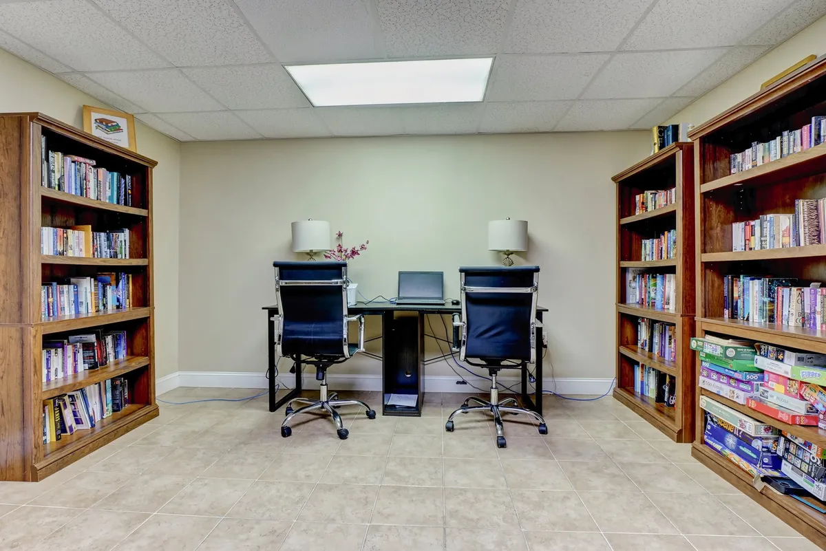 A serene business center featuring bookshelves lined with books, two comfortable black chairs, and a desk with a computer. The environment is quiet and conducive to remote work or study.