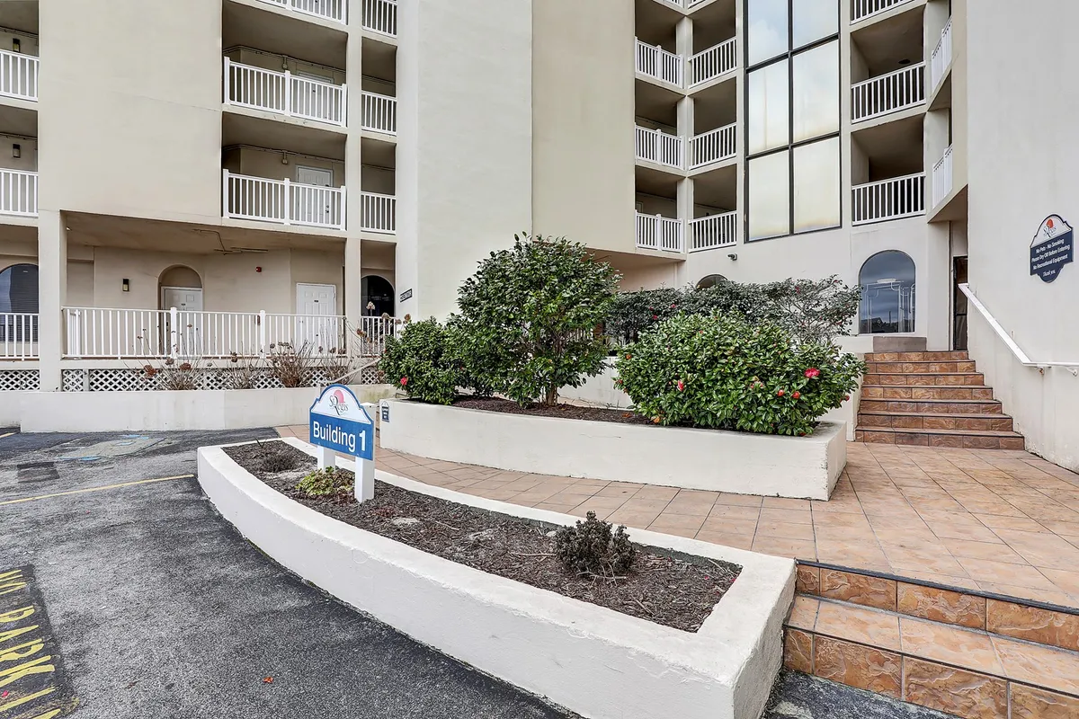 The image displays the exterior entrance of the building, featuring prominent building signage, a landscaped area, and a welcoming staircase leading up to the main doors. The scene is bathed in natural light, inviting guests into the property while glimpses of the surrounding greenery set a relaxed, vacation-ready mood.
