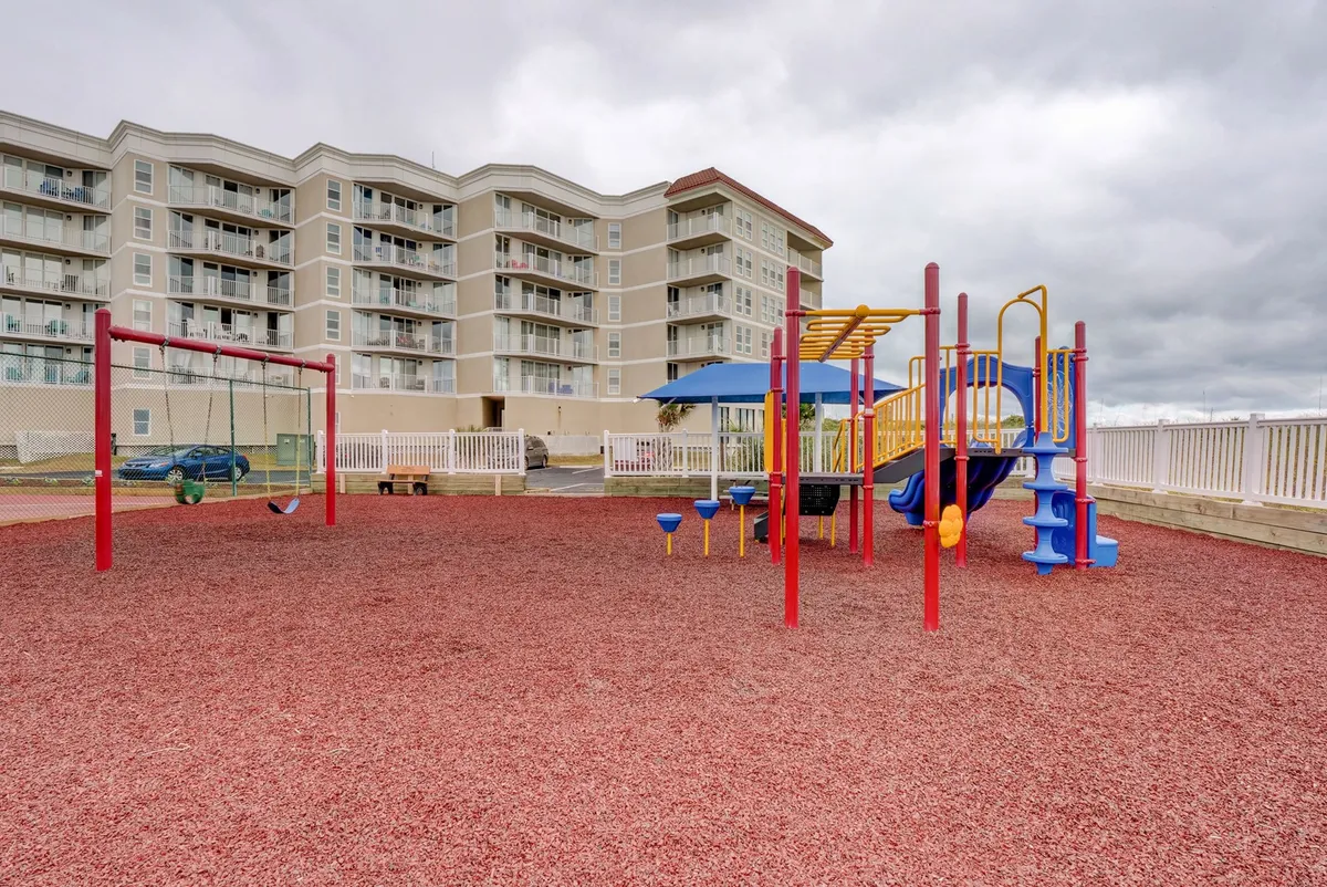 A cheerful playground set on a sand area, equipped with swings and slides, with a building visible in the background. Children play energetically while parents and families gather nearby.