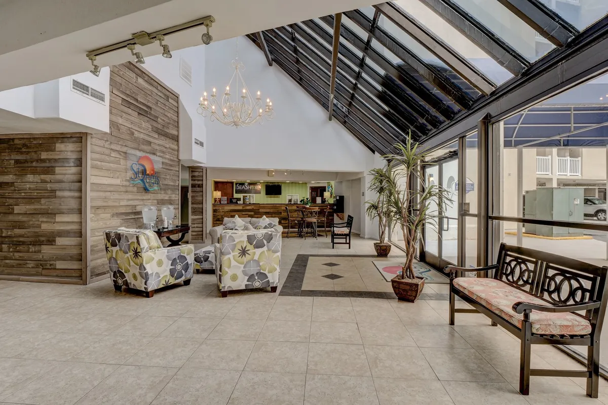 A spacious, light-filled reception area with a chandelier overhead, a welcoming reception desk, and lounge seating surrounded by large windows. The natural light and open feel offer a fitting first impression for guests arriving to blend work and leisure.