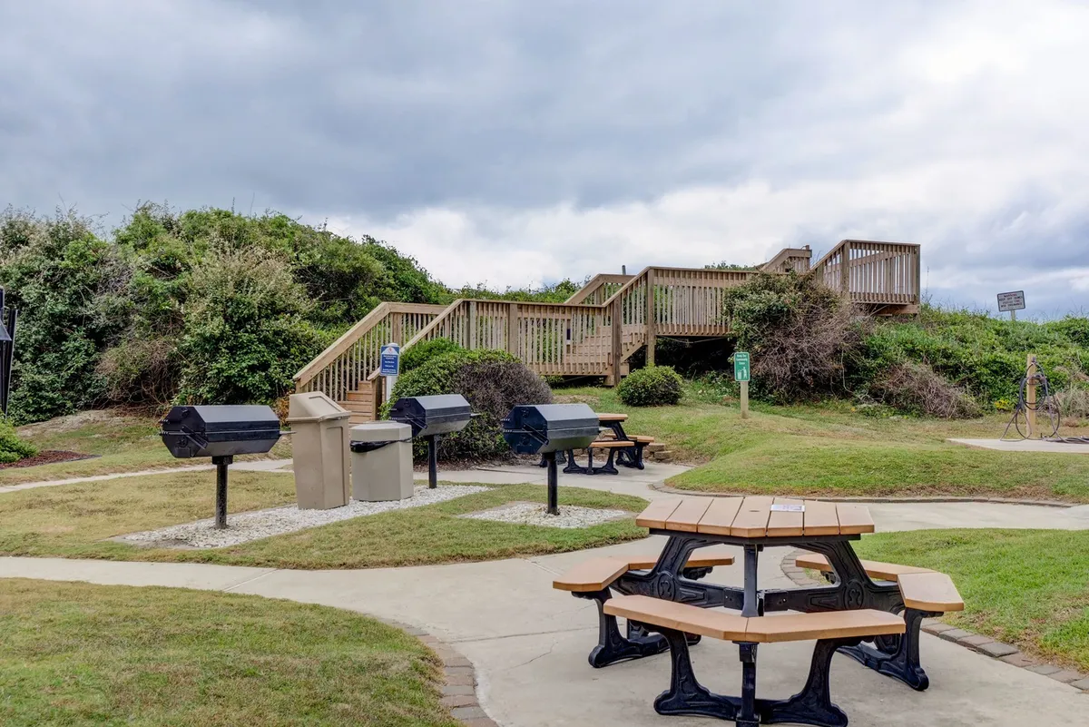 Outdoor picnic tables paired with barbecue grills and a surrounding relaxed communal setting. The scene is designed for socializing and enjoying meals al fresco, ideal for casual gatherings or family cookouts.