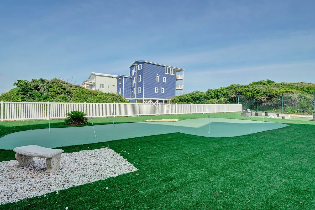 The image showcases a green putting green surrounded by a white fence and a blue building, with a stone bench inviting guests to relax. The open outdoor area is ideal for enjoying leisurely golf practice or socializing under sunny skies.
