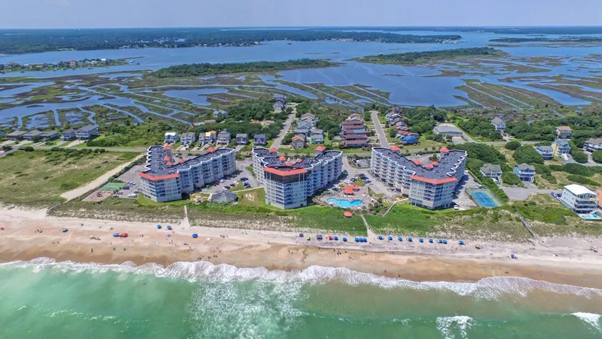 The hero image showcases a modern beachfront condominium complex set against the backdrop of North Topsail Beach, with a sparkling, covered saltwater pool in the foreground, sweeping views of the ocean, and tranquil marshland stretching into the horizon. The scene captures sunset light reflecting off the water and inviting lounge spaces around the pool, evoking a sense of relaxation and coastal beauty.