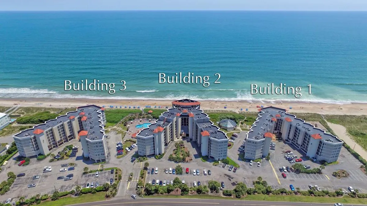 A beachfront property with a sparkling swimming pool, sun loungers, and three buildings facing the ocean. The pool area is lively but relaxed, with families enjoying the water and the proximity to the beach.